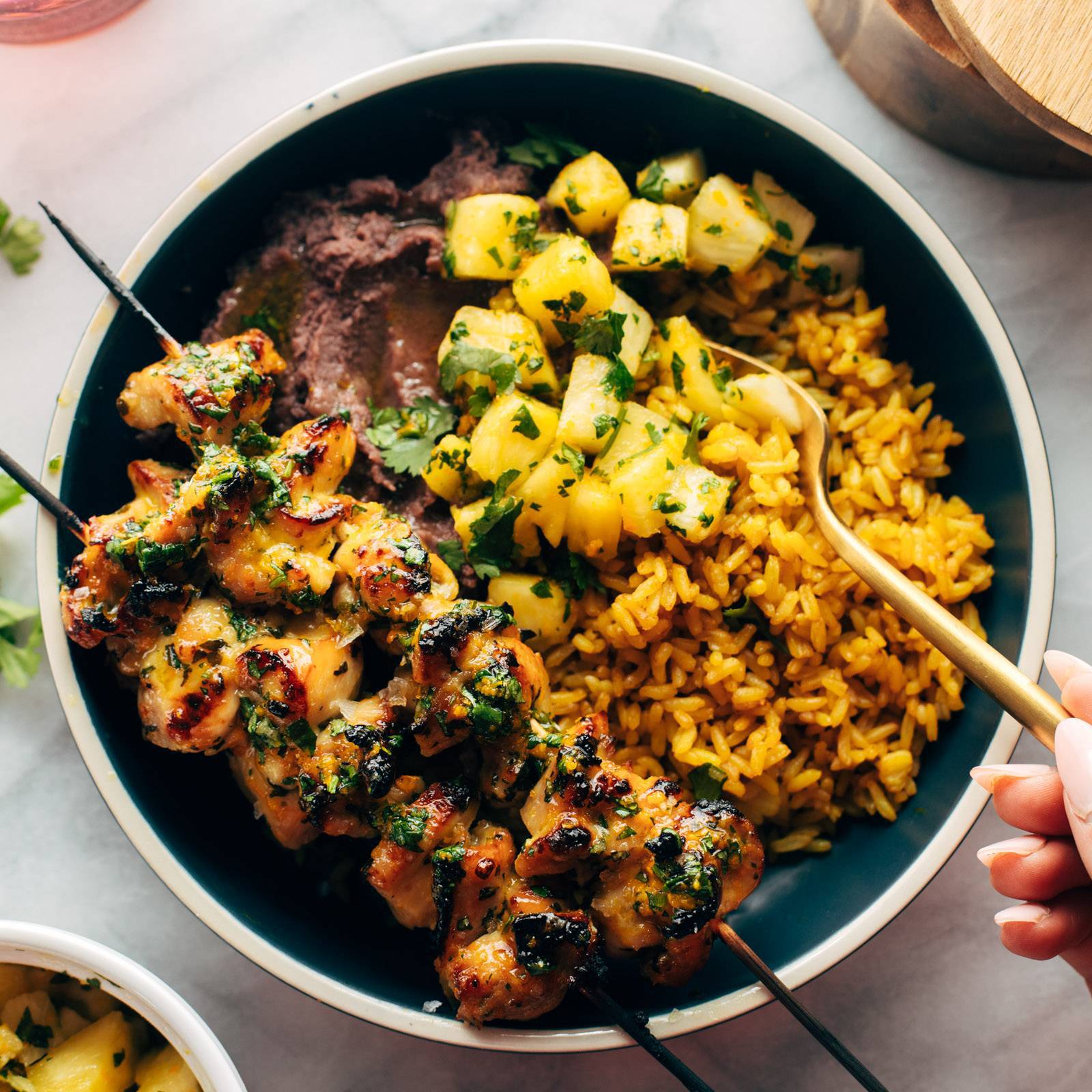 Cilantro orange chicken on skewers over a bowl of rice, pineapple, and black beans.